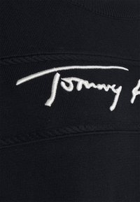 Tessuto nero con una texture a costine, caratterizzato da un logo firmato "Tommy" ricamato in bianco. Cuciture dettagliate accentuano le cuciture.