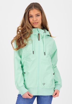 Jeune femme portant une veste à capuche zippée couleur menthe avec des cordons, posant devant un fond blanc uni.