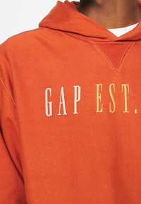 Felpa arancione con logo ricamato bianco e giallo "GAP EST.". Tessuto morbido con tasca a marsupio e cappuccio con coulisse regolabile.