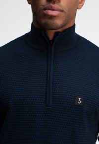 Navyblauwe ritstrui met een textuur breipatroon, hoge kraag en een klein zwart logo-opdruk op de borst.