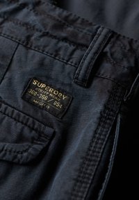 Tejido de un azul marino oscuro con una textura suave, que presenta una etiqueta rectangular negra con texto en blanco y dorado en el área del bolsillo trasero.