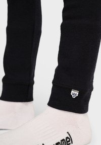 Svarta, tight passformade träningsbyxor i mjukt material, med en fållad manschett och en liten vit logotyp på nederdelen av benet. Vit strumpa synlig.