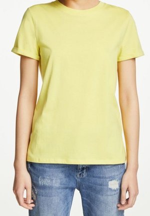 Persoon die een effen lichtgeel T-shirt met korte mouwen en blauwe versleten spijkerbroek draagt, staande tegen een witte achtergrond.