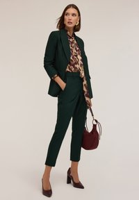 Groen op maat gemaakt pak met slim-fit broek en een bijpassende blazer. Gelaagd met een gebloemd blouse, houdt een bordeauxrode handtas vast en draagt hakken.