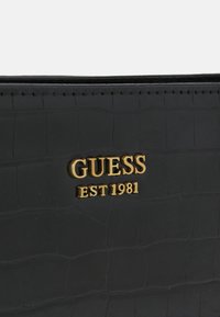 Portefeuille noir en simili-crocodile avec le logo doré "GUESS EST 1981". Présente une surface texturée et des coutures soignées le long des bords.