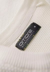 Tela blanca texturizada con un patrón tejido, que presenta una etiqueta negra que muestra "Ombre Quintessence" y la talla "M" con detalles en verde.