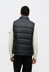 Μαύρο επενδεδυμένο γιλέκο με ψηλό γιακά, με ραμμένα τμήματα και λεία υφή. Φοριέται πάνω από ένα λευκό, υφασμάτινο πουλόβερ.