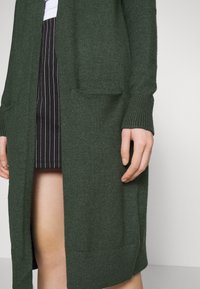 Lång, mörkgrön kofta i strukturerad stickad kvalitet, med två framfickor och ribbade ärmslut, bärs över en randig kjol.