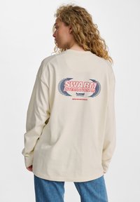 Cremfarbendes Langarmshirt mit Rückendruck "SWARM INTERNATIONAL" in Rot und Blau, mit lockerer Passform und weichem Stoff.