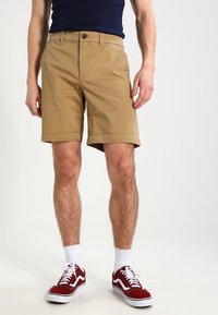 Shorts beiges à coupe droite, poches avant et ourlet retroussé, portés avec des baskets rouges et blanches et des chaussettes blanches.