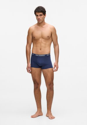 Čovjek bez majice stoji bos, nosi tamnopoplasnate bokserice s bijelim pojasom na kojem piše "KARL LAGERFELD", na bijeloj pozadini.