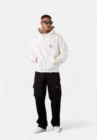 White hooded sweatshirt with a front pocket and graphic detail, paired with black cargo pants featuring patch pockets and white sneakers.