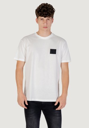 Camiseta de algodón blanca con un bolsillo cuadrado negro que lleva un logo. Mangas cortas y un corte relajado. Combinada con jeans oscuros.