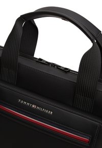 Κοντινό πλάνο μαύρης τσάντας Tommy Hilfiger με υφασμάτινες λαβές, φερμουάρ και λογότυπο με κόκκινες, λευκές και μπλε ρίγες.