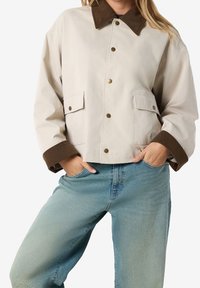 Person iført en beige jakke med brunt krave og manchetter, hænderne i lommerne, parret med lyseblå jeans mod en ensfarvet baggrund.