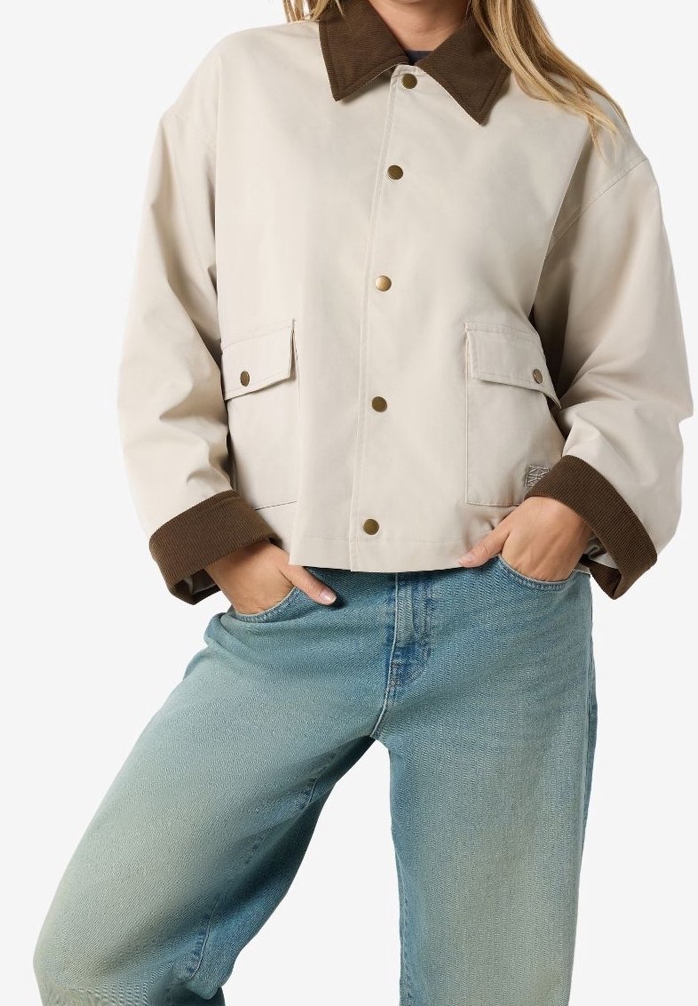 Person iført en beige jakke med brunt krave og manchetter, hænderne i lommerne, parret med lyseblå jeans mod en ensfarvet baggrund.