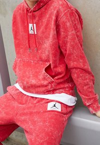 Punainen tikkitakki, jossa on etupuolella tasku ja logopatch; yhdistettynä vastaaviin collegehousuihin, joissa on logopatch reidessä.