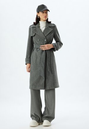 Cappotto trench grigio in lana con bottoni doppi, vita con cintura e ampi revers, abbinato a pantaloni grigi a gamba larga e un cappello.