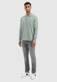 Hellgrünes Langarm-Polo mit zwei Knöpfen, kombiniert mit grauen Jeans und weißen Sneakers mit blauen Akzenten. Glatte Stofftextur.