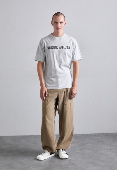 Gray cotton t-shirt with "Moschino Couture!" printed in black, paired with loose beige cargo pants and white sneakers on a gray background.