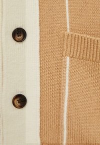 Cardigan en tricot aux couleurs beige clair et camel, avec deux boutons marron foncé et des poches texturées ornées d'une ligne blanche.