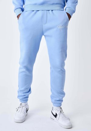 Persona che indossa pantaloni della tuta azzurri con le mani nelle tasche e sneakers Nike bianche con swoosh nero.