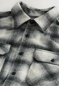 Grau- und schwarz kariertes Flanellhemd mit Kragen, ausgestattet mit zwei aufgesetzten Brusttaschen mit Knöpfen und schwarzen Kunststoffknöpfen. Weiche Textur.