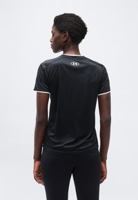 Γυ ναίκα με κοντά κατσαρά μαλλιά που φοράει μαύρο μπλουζάκι και κολάν Under Armour, εμφανιζόμενη από πίσω σε απλό ανοιχτό φόντο.