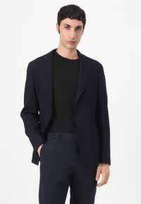 Blazer navy con solapas de muesca, cierre de un solo botón y dos bolsillos frontales, llevado sobre una camiseta negra y combinado con pantalones oscuros.