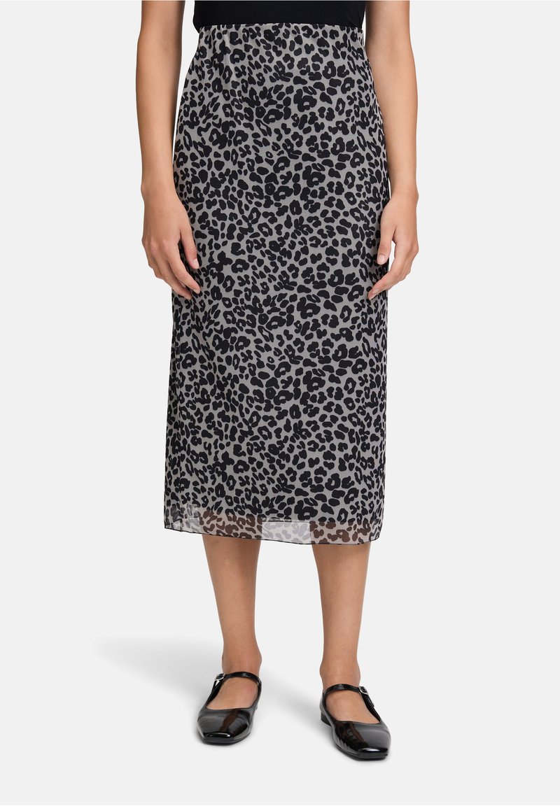 Frau in einem schwarz-grauen Leopardenmuster-Midirock, schwarzem Oberteil und schwarzen Mary-Jane-Schuhen, die vor einem schlichten Hintergrund steht.