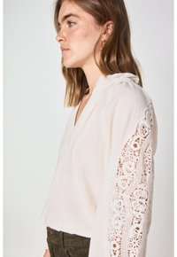 Blusa crema con dettagli in pizzo sulle maniche, caratterizzata da un colletto a V, bottoni lungo la parte anteriore e un tessuto morbido e testurizzato.