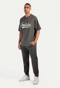 Grijs oversized t-shirt met het "Smilodox"-logo in het wit, gecombineerd met bijpassende grijze joggingbroek en witte sneakers. Zachte stof, ontspannen pasvorm.