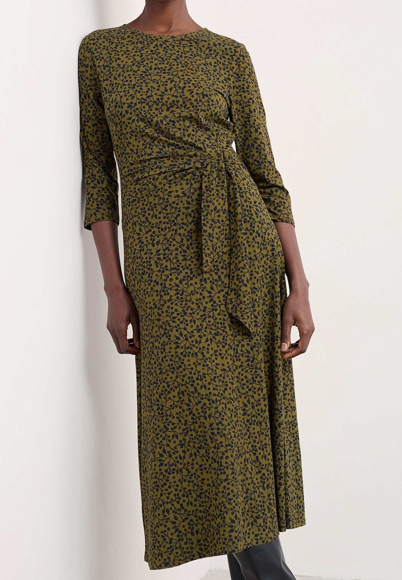 Personne à la peau foncée portant une robe vert olive avec un motif de feuilles noires, manches trois-quarts, nœud latéral, debout contre un mur blanc uni.