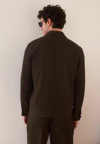 Chaqueta marrón a medida con una textura suave, costura trasera única y puños abotonados. Llevada con pantalones a juego, exhibida contra un fondo neutro.