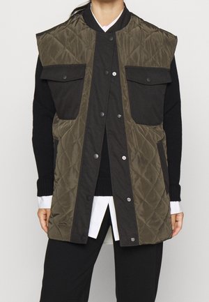 Osoba w luźnej, bezrękawnikowej, oliwkowozielonej pikowanej kamizelce z czarnymi kieszeniami z patkami, nałożonej na czarny sweter i białą koszulę.