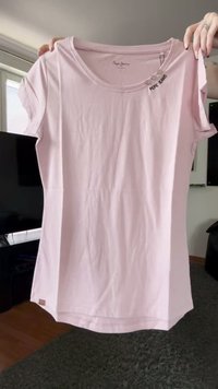 Manos sosteniendo una camiseta de Pepe Jeans de manga corta, color rosa claro, con escote redondo y detalles de botón cerca del collar.