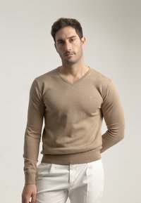 Suéter beige de manga larga con cuello en V, confeccionado en una tela suave, con un diseño ajustado y puños acanalados, combinado con pantalones blancos.