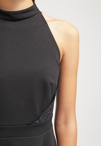 Černé šaty s halterem, vyrobené z hladké látky s texturovanými krajkovými bočními panely a přiléhavým střihem.