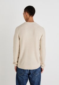 Beige gebreide trui met een gestructureerd oppervlak, ribbelboord bij de ronde halslijn en lange mouwen, gedragen met blauwe spijkerbroek. Beeld vanaf de achterkant.