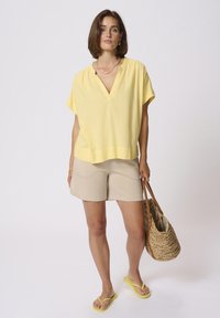 Blouse jaune à manches courtes avec un décolleté en V, associée à un short beige. Le mannequin porte un sac en paille tissé et des tongs jaunes.