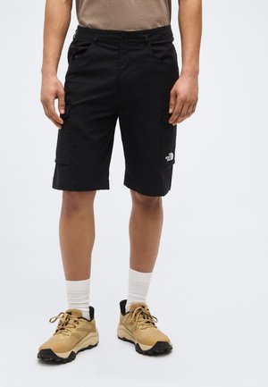 Uomo che indossa pantaloncini cargo neri con tasche e scarpe da trekking color sabbia con calzini bianchi, in piedi su uno sfondo uniforme.