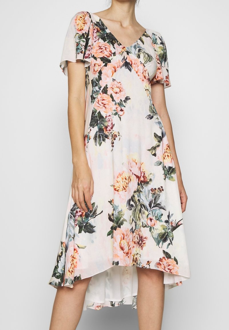 Femme portant une robe blanche à fleurs roses et oranges, avec des manches courtes volantées et un ourlet haut-bas.