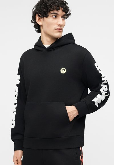 Ung mann iført svart hettegenser med lite poteavtrykk-logo på brystet og teksten "BARREL" i hvitt ned begge ermene, hånden i frontlommen.