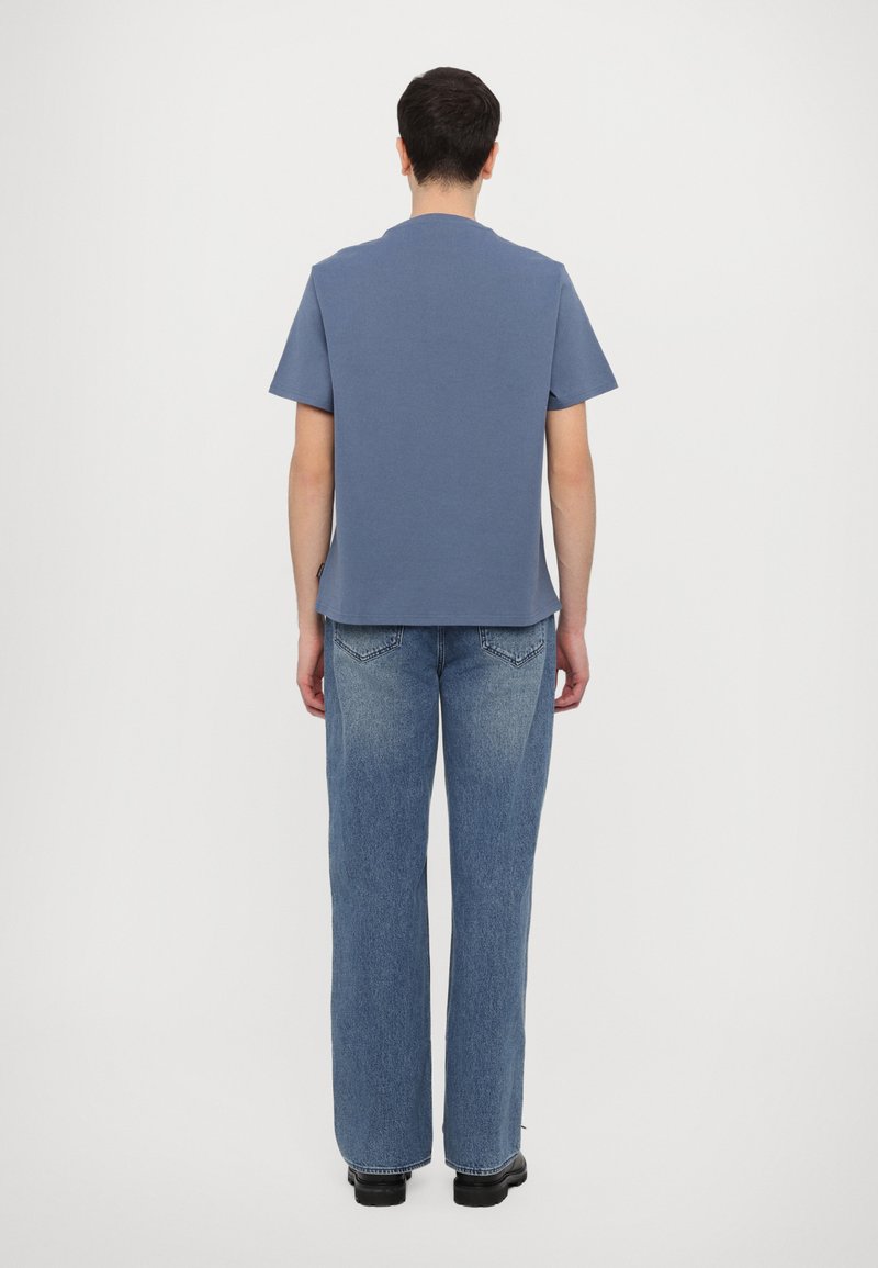 Hellblaues Baumwoll-T-Shirt mit kurzen Ärmeln und geradem Saum, kombiniert mit hellblauen geraden Jeans und schwarzen Schuhen, von hinten betrachtet.