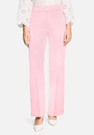 Pantalon rose évasé avec une texture lisse et une taille haute, associé à des chaussures à talons blanches et un chemisier blanc en broderie anglaise.