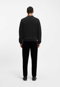 Felpa nera a maglia con colletto, abbinata a pantaloni neri in corduroy. Il modello indossa scarpe nere e sta girato di spalle rispetto alla fotocamera.
