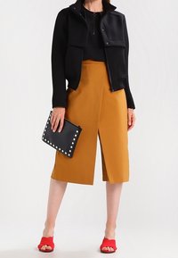 Giacca nera, gonna asimmetrica senape, sandali rossi e pochette nera con borchie. Materiali morbidi e contrasti di colore definiti.