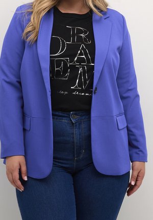 Žena v královsky modrém saku přes černé tričko s nápisem "SNY" ve velkých stříbrných písmenech a tmavě modrých džínách s vysokým pasem.