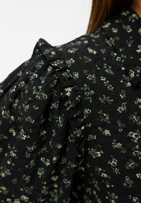 Blusa nera a fiori con motivi verdi e bianchi, dotata di spalle arricciate e tessuto testurizzato. Inquadratura ravvicinata.