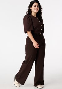 Bruine top met pofmouwen en gouden knopen, gecombineerd met hoge, rechte bruine broek en witte slip-on schoenen met patroon.